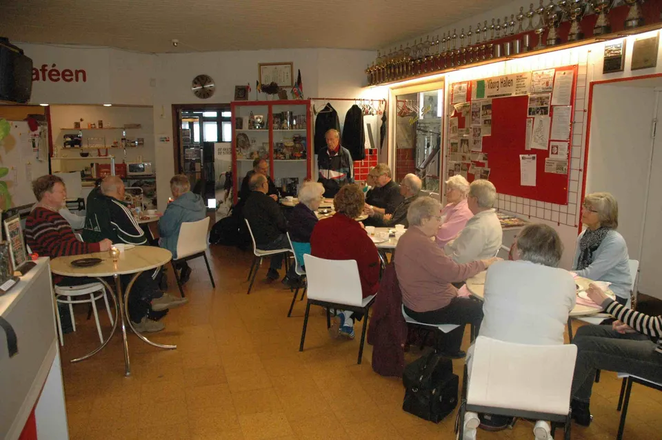 Seniorer sidder ved bordene i caféen med billeder og pokaler på væggene